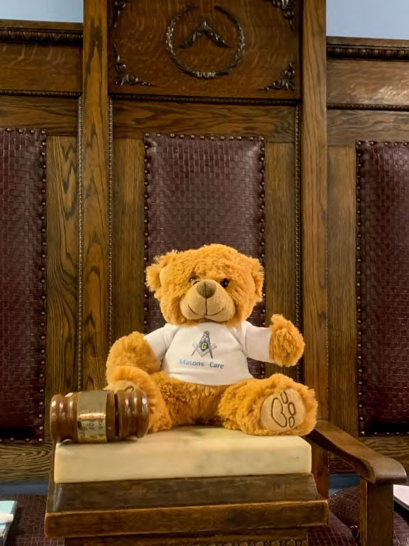 A Teddy Bear is seen sitting on a gavels sound block with the Worshipful Masters gavel by his right side. He is in front of the chair in the East. The Teddy Bear is wearing a shirt saying "Masons Care".